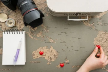 Uma imagem de vista superior mostra um mapa mundial antigo e de cor cinza sobre uma superfície de madeira. No canto superior esquerdo, há uma câmera reflex monobjetiva preta com uma lente 24-70 mm acoplada, ao lado de uma bússola dourada aberta. No canto inferior esquerdo, há um caderno de espiral aberto com páginas em branco, acompanhado por uma caneta roxa. Ao longo da borda superior, há uma mala de viagem de cor bege claro fechada. Sobre o mapa, no continente australiano, estão dois corações vermelhos, e no sudeste, perto da Nova Zelândia, está um terceiro coração vermelho. Uma mão direita feminina aparece no canto inferior direito, segurando um pequeno coração vermelho sobre a América do Sul. A imagem é bem iluminada e foca nos detalhes dos objetos.