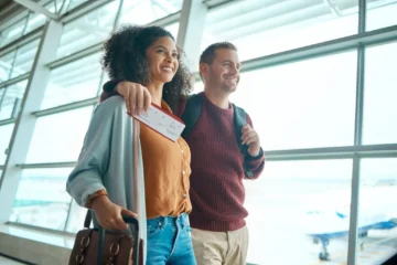 Um casal sorridente caminha abraçado pelo saguão bem iluminado de um aeroporto, em frente a grandes janelas de vidro que revelam uma aeronave ao fundo. À esquerda, uma mulher negra com cabelos cacheados usa uma jaqueta clara sobre uma blusa mostarda e calça jeans, segurando uma passagem de avião na mão direita e uma mala de rodinhas com a outra. À direita, um homem branco com cabelo curto e barba rala veste um suéter de tricô marrom, calça cáqui e carrega uma mochila nas costas, com o braço direito apoiado nos ombros da companheira. Ambos olham para frente com otimismo, capturando a expectativa do início de um intercâmbio.